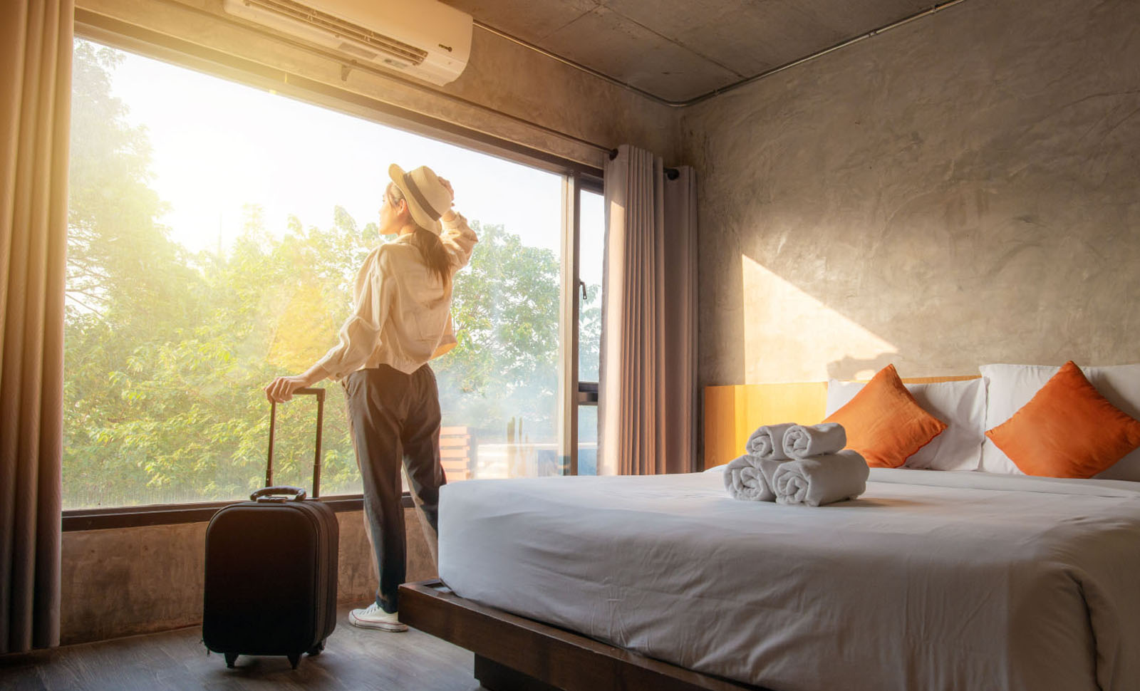 Woman looking out the window of her hotel room holding her luggage in one hand.