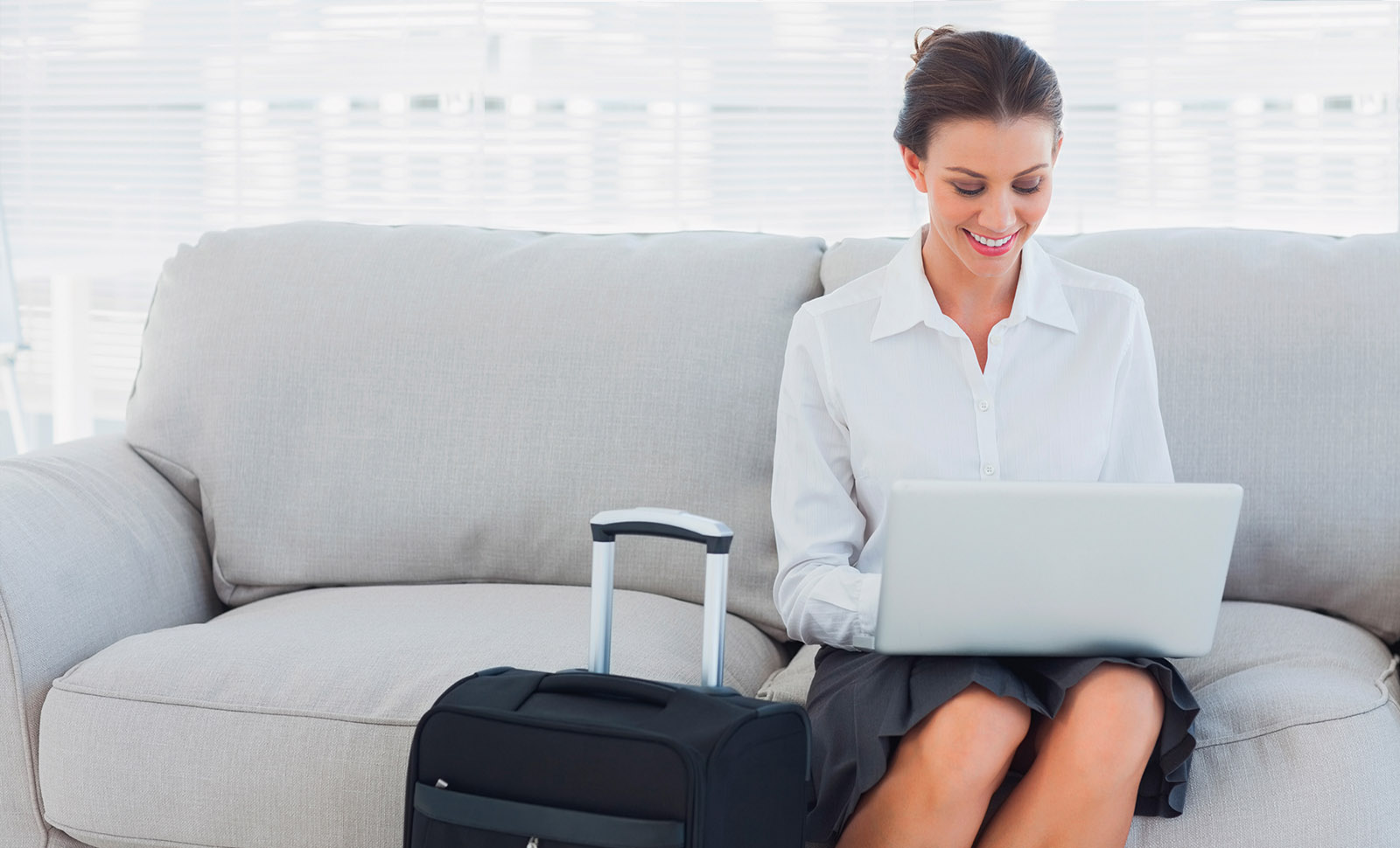 woman sitting on couch in hotel room working on her laptop with her suitcase next to her